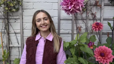 Hannah is a woman in her early 30s. She has long blonde hair and is smiling at the camera. She's pictured against a white brick wall and has tall dahlias growing either side of her. She is wearing a pink collared shirt with a dark red waistcoat. 