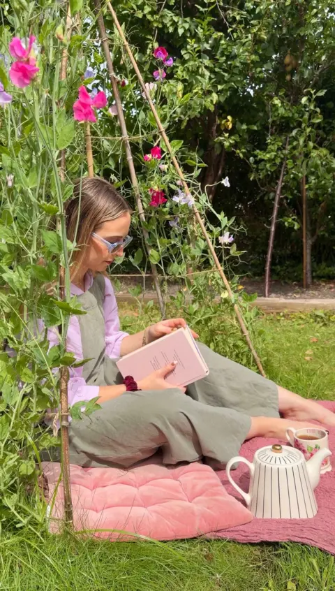 Hannah Matthews Hannah sitting on a mat in her back garden, reading a book, surrounded by plants