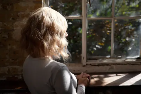 Laura sits by a sunlit window, her hands clasped as warm light catches her hair.