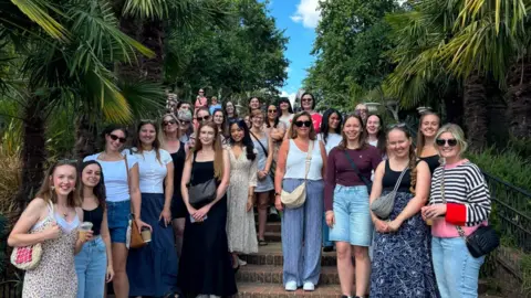 Georgia Featherstone This photograph includes more than 20 people on the steps of a garden. They're all various ages and heights. There are trees lining the steps.
