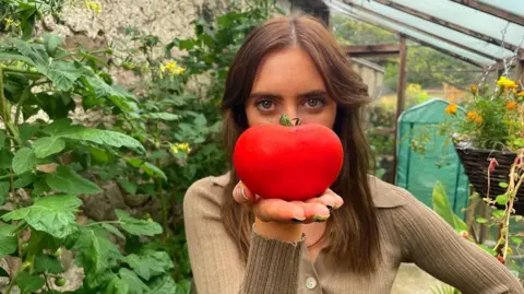 Naomi Saunders Naomi is pictured her in a greenhouse against a backdrop of flowers. She's holding a large red tomato in front of her face, placed on her palm. She has long brown hair and is wearing a knit green top.